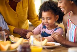 Família preparando alimentação saudável para crianças com frutas, incluindo bananas e maçãs, durante uma refeição ensolarada