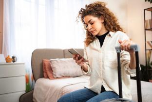A imagem mostra uma mulher de cabelos cacheados em um quarto bem iluminado, sentada na beira da cama e segurando uma mala de rodinhas. Ela está concentrada olhando para a tela de um smartphone, sugerindo a organização de uma viagem, reserva de hospedagem ou verificação de bilhetes aéreos. O cenário transmite uma atmosfera de preparação para viagem, conveniência digital e planejamento de turismo através de aplicativos móveis.