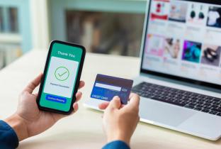 A imagem em close-up destaca as mãos de um usuário segurando um smartphone que exibe uma interface de confirmação de pagamento (wallet digital), enquanto a outra mão segura um cartão de crédito. Ao fundo, um laptop exibe um site de e-commerce, reforçando o conceito de compras seguras na internet. A cena comunica agilidade, tecnologia de pagamento por aproximação ou carteira digital e a integração entre dispositivos para uma experiência de consumo fluida e moderna.