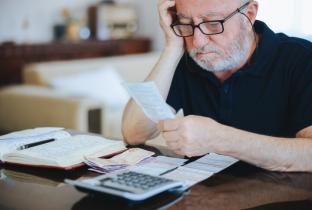 A imagem mostra um senhor de óculos sentado à mesa, com a mão na cabeça, demonstrando estresse ou dificuldade financeira. À sua frente, há diversos boletos, papéis de cobrança, um caderno aberto e uma calculadora. A cena retrata a gestão do orçamento doméstico, o impacto do aumento de custos ou a organização de dívidas, com foco na tensão emocional causada pelo planejamento financeiro na terceira idade.