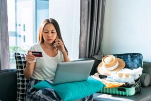 A imagem mostra uma jovem de expressão tensa e confusa sentada em um sofá, conversando ao smartphone enquanto segura um cartão de crédito. Em seu colo, há um notebook apoiado em uma almofada azul. Ao seu lado, uma mala de viagem verde está aberta e arrumada com roupas, óculos de sol e um chapéu. A cena retrata um momento de frustração ou urgência relacionado a serviços de turismo, como a necessidade de contestar uma cobrança, resolver um imprevisto de última hora ou cancelar uma reserva no Airbnb.