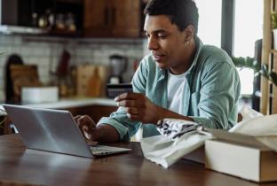 A imagem mostra um homem negro em um ambiente doméstico (cozinha/sala), concentrado enquanto digita informações em um laptop. Ele segura um cartão de crédito, indicando uma transação financeira em andamento. À sua frente, há uma caixa de encomenda aberta com papéis de embalagem, o que sugere que ele pode estar tanto finalizando uma nova compra quanto inserindo dados para solicitar um reembolso de um produto recém-chegado. A cena transmite praticidade e o uso de tecnologia para resolver questões de consumo n
