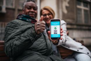 A imagem mostra um senhor negro de casaco verde e cachecol em primeiro plano, sorrindo e gesticulando com o polegar levantado. Ao seu lado, uma mulher desfocada ao fundo segura e aponta um smartphone em direção à câmera. A tela do celular exibe um documento digital azul com um código QR destacado. A cena se passa em um ambiente externo urbano e retrata o uso de tecnologia na terceira idade, acessibilidade, direitos da pessoa idosa e a facilidade de portar carteirinhas ou comprovantes de identificação
