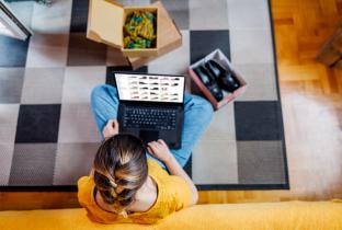 A imagem, capturada de um ângulo superior (top-down), mostra uma pessoa com um laptop no colo navegando em um site de vendas. Ao redor dela, sobre um tapete geométrico, há uma caixa de papelão aberta com produtos e um tablet, simbolizando a conveniência do consumo digital e a experiência de receber encomendas em casa. O cenário transmite uma atmosfera de conforto doméstico e a facilidade de comparar preços e produtos através de plataformas de marketplace.