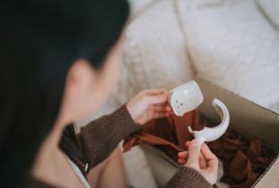 A imagem mostra, em primeiro plano e com foco seletivo, as mãos de uma pessoa segurando os cacos de uma peça de cerâmica ou porcelana branca quebrada. O objeto está sendo removido de uma caixa de transporte com papel de proteção pardo. A foto enfatiza a falha na logística de entrega e o dano material, sendo ideal para ilustrar conteúdos sobre avarias no transporte, direitos de troca e embalagem inadequada no e-commerce.