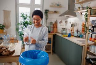 A imagem mostra uma mulher negra em uma cozinha organizada realizando a separação de resíduos. Ela segura uma garrafa de plástico transparente, comprimindo-a para reduzir o volume antes do descarte. O ambiente é iluminado, com cesto de lixo destacado em primeiro plano e elementos de decoração sustentável ao fundo, como plantas e prateleiras de madeira.