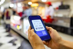 A imagem apresenta um close-up das mãos de uma pessoa segurando um celular que mostra um e-ticket (ingresso eletrônico) azul. Na tela, são visíveis informações como horário, assento e os códigos de validação. O fundo está desfocado (bokeh), sugerindo o saguão de um cinema, teatro ou arena de eventos, destacando a praticidade do uso de bilhetes digitais e a tecnologia de acesso via dispositivos móveis.